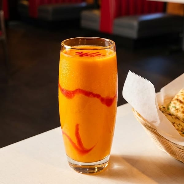 Orange mango lassi with red swirl, next to a basket of naan bread on a white table.