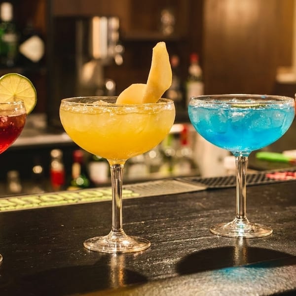 Four colorful cocktails, red, yellow, blue, and red, lined up on a dark bar counter.